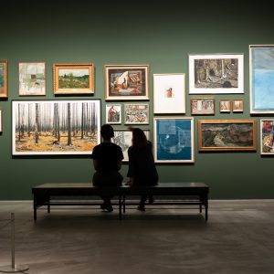 målningar och fotografier hänger tätt på grön vägg två personer sitter på bänk och iakttar målningarna