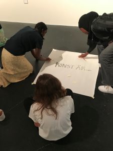 tre ungdomar sitter på golvet och tittar på ett stort vitt papper med texten konst är...